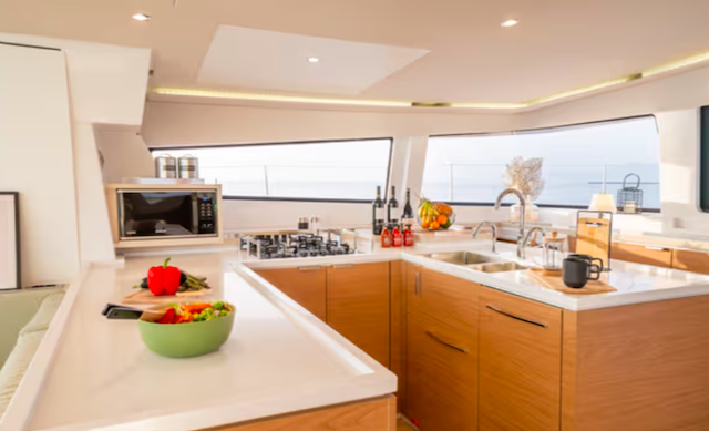 Modern yacht kitchen with wooden cabinets, a microwave, a sink, and a stove. A bowl of vegetables, including a red pepper, is on the counter. Bottles and oranges are near the sink. Large windows provide a sea view.