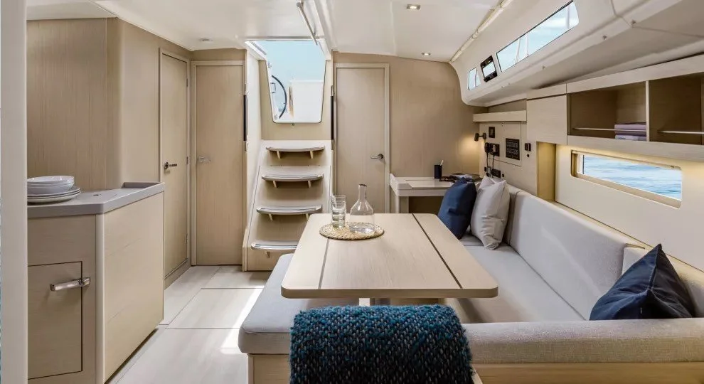 Interior of a modern yacht showing a dining area with a wooden table, cushioned seating with pillows, and a small kitchen area to the left. The space features light wood finishes, a staircase leading to the deck, and windows providing natural light.