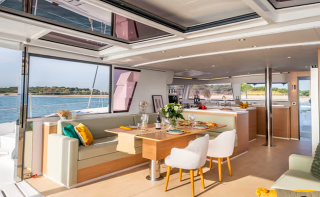 Interior of a yacht with a dining area and seating, featuring a table set for a meal, cushioned benches, and modern decor. Large windows offer a view of the water and shoreline.
