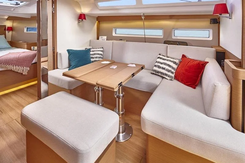 Interior view of a yacht cabin with a cozy seating area featuring cream-colored cushions and a wooden table. Decorative pillows in red, blue, and black-and-white patterns add contrast. The cabin has modern minimalist design elements and natural lighting from small windows.