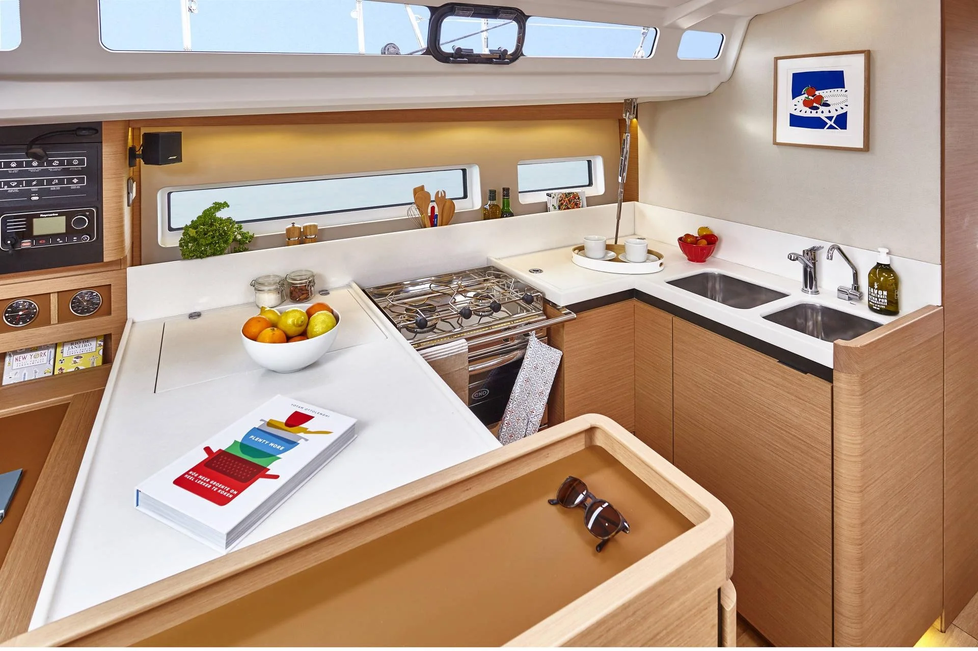 Interior of a yacht kitchen with wooden cabinets, white countertops, a stove, sink, and window. Includes a fruit bowl, books, a framed picture, and kitchen utensils.