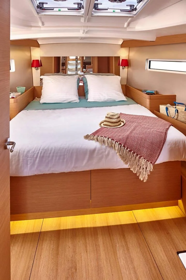 Cozy boat cabin interior with a neatly made bed, red lamps, a straw hat, and a fringed red blanket. Wooden floors and cabinetry, with a window and a skylight. Items on the shelves include a striped bag and a patterned mug.