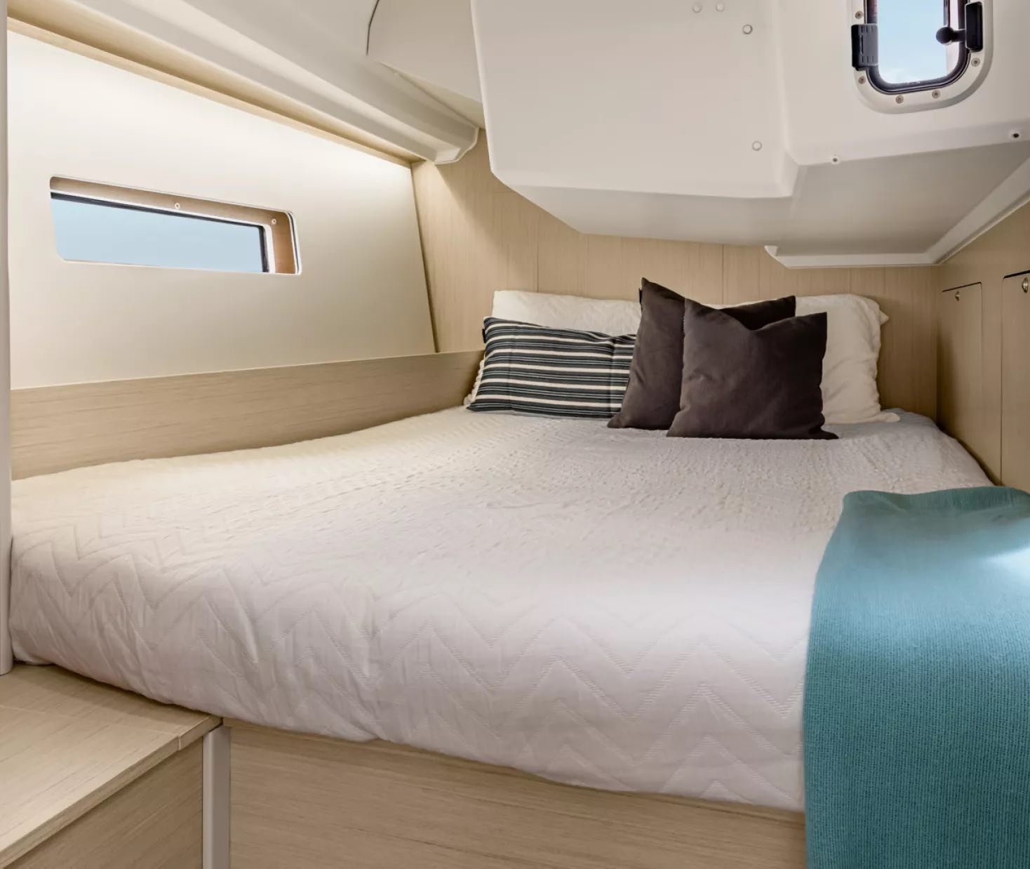 Small, neatly made bed in a yacht cabin with a window, white bedding, gray and striped pillows, and a blue throw blanket.