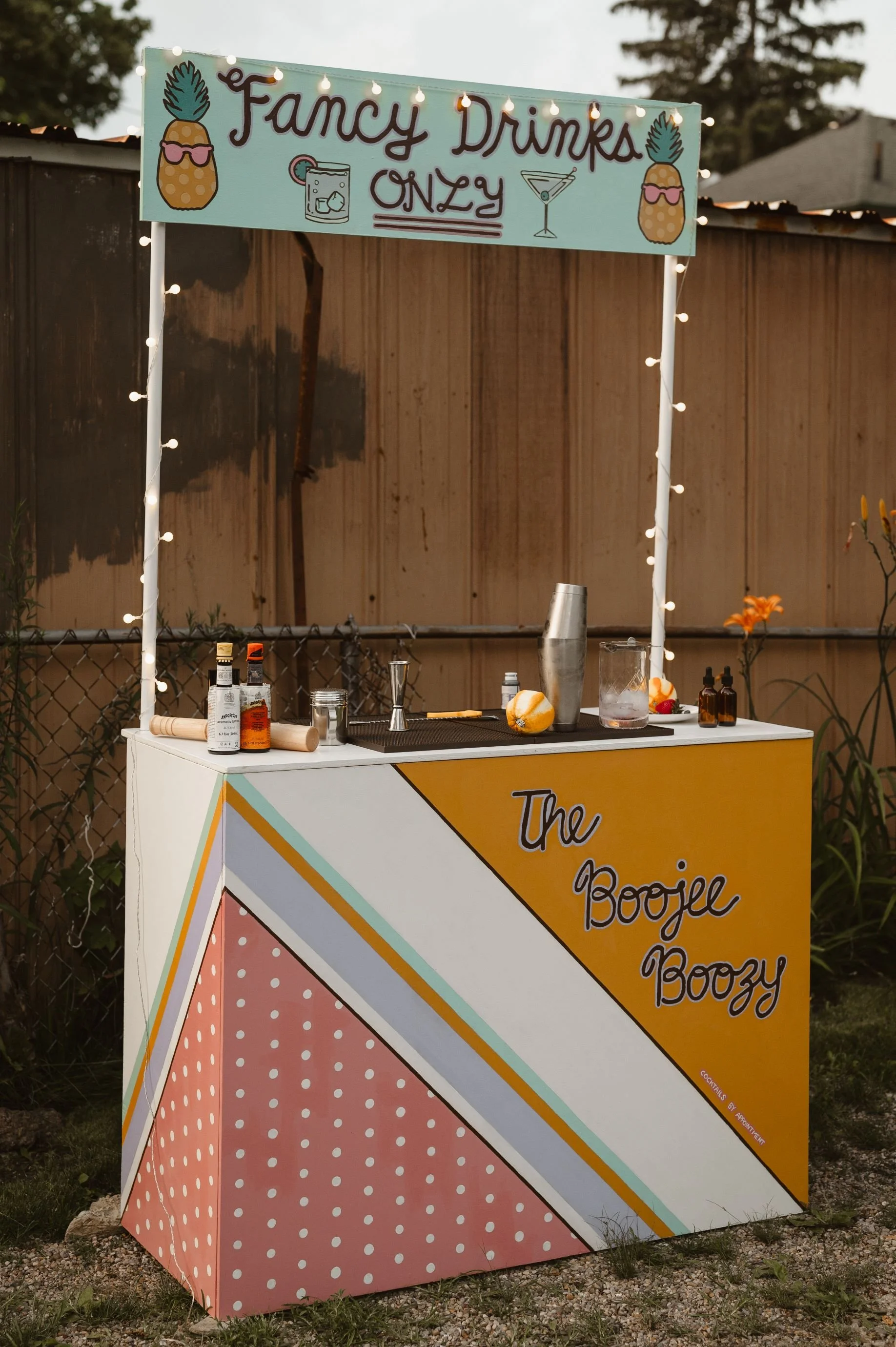 A portable bar setup titled 'The Boozie Boozy' with a colorful painted front panel, with bottles, a cocktail shaker, and bar tools on top. A sign above reads 'Fancy Drinks Only' with drawings of pineapples wearing sunglasses and cocktail glasses, and is decorated with string lights.