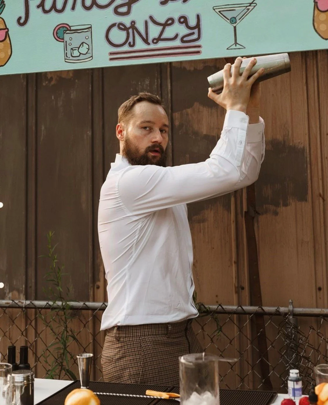 A man in a white shirt and plaid pants is shaking a cocktail shaker above his head, standing behind a bar counter with bartending tools and ingredients, with a wooden fence and a colorful sign above him.