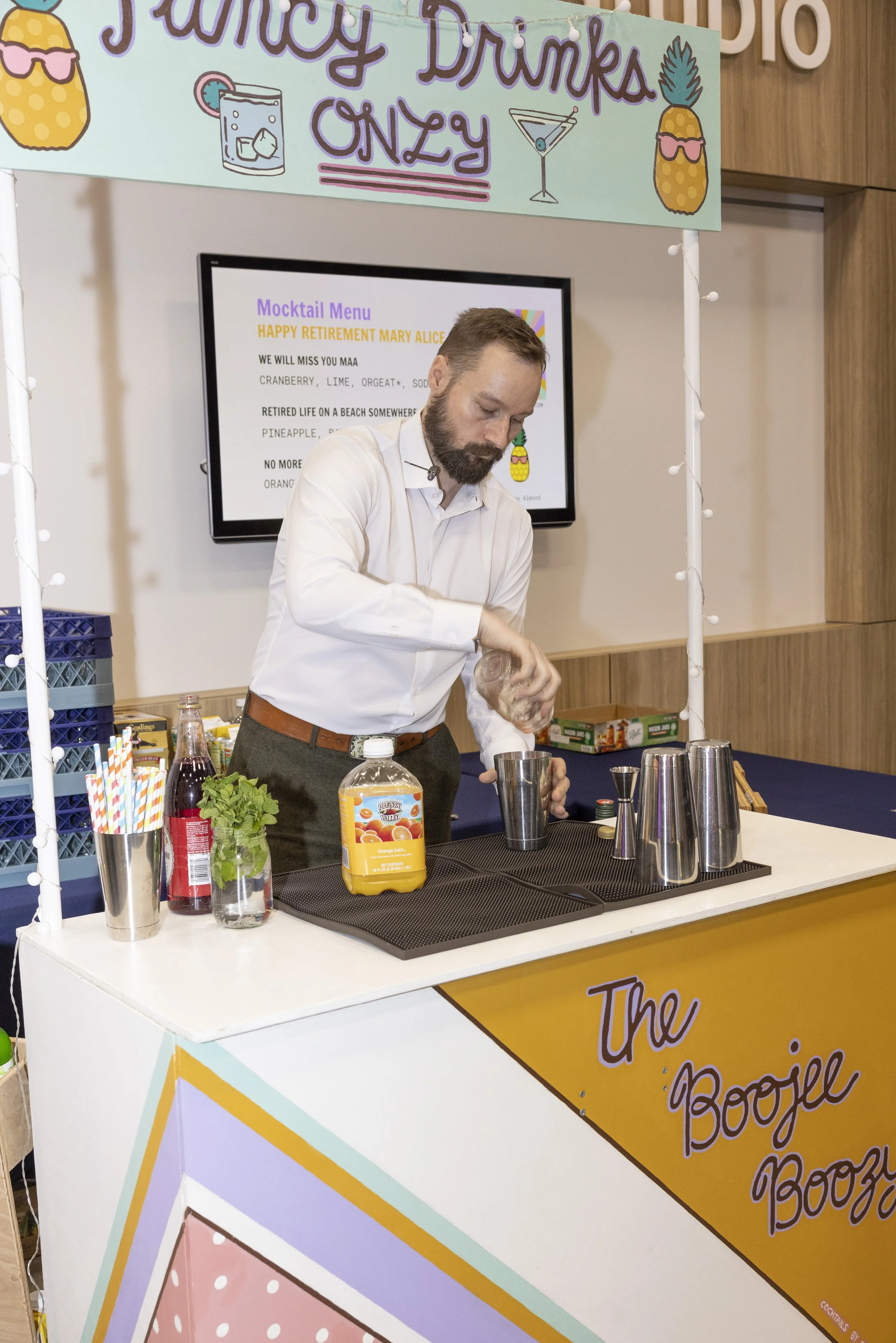 A man preparing a cocktail at a juice bar stand labeled "The Boozy Booz". The stand has a colorful sign overhead that says "Juicy Drinks Only" with images of tropical fruits and drinks. The man is pouring a liquid into a metal shaker, with various cocktail shakers, juice bottles, and garnishing ingredients on the counter.