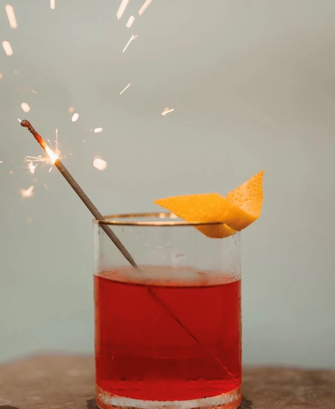 A red cocktail in a clear glass with an orange garnish, a lit sparkler in the drink, and a lemon twist on the rim, against a plain background.