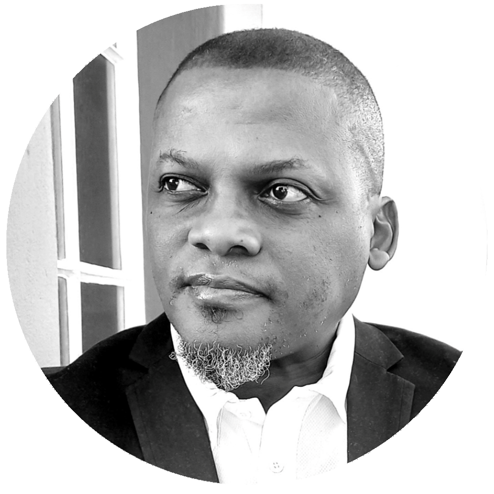 Black and white close-up portrait of a man with short hair, a goatee, wearing a white shirt and dark blazer, looking to the side with a thoughtful expression.