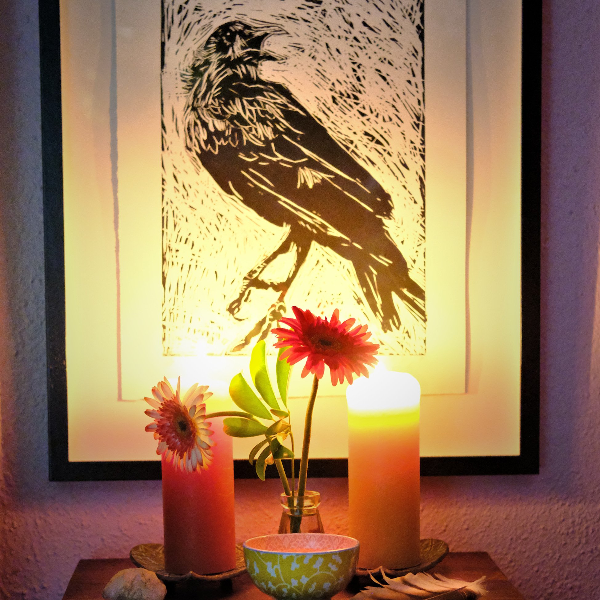 A framed black and white bird illustration hanging on a wall, with lit candles and a vase of flowers and a bowl on a table in front of it.
