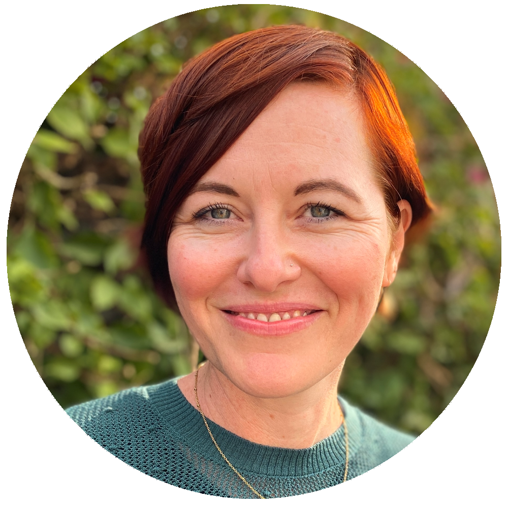 A woman with short, reddish-brown hair smiling outdoors with green foliage in the background.
