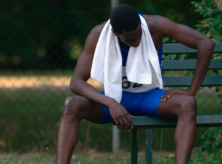 Male athlete in blue sportswear and a white towel sitting on a park bench, appearing exhausted.