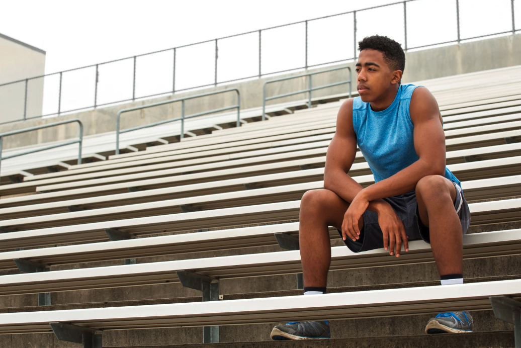 A young man in a blue athletic tank top and black shorts sitting alone on empty stadium bleachers, looking thoughtfully into the distance.