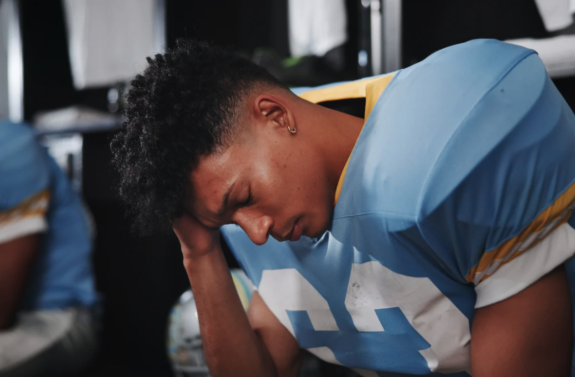 A young football player in a blue and white jersey appears distressed, with his head bowed and resting on his hand in a locker room.