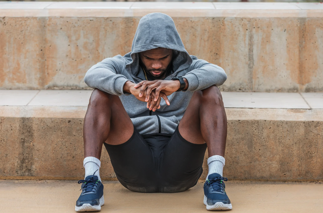 A young man in athletic attire, wearing a gray hoodie and black shorts, sitting on concrete steps with his head down and hands clasped.