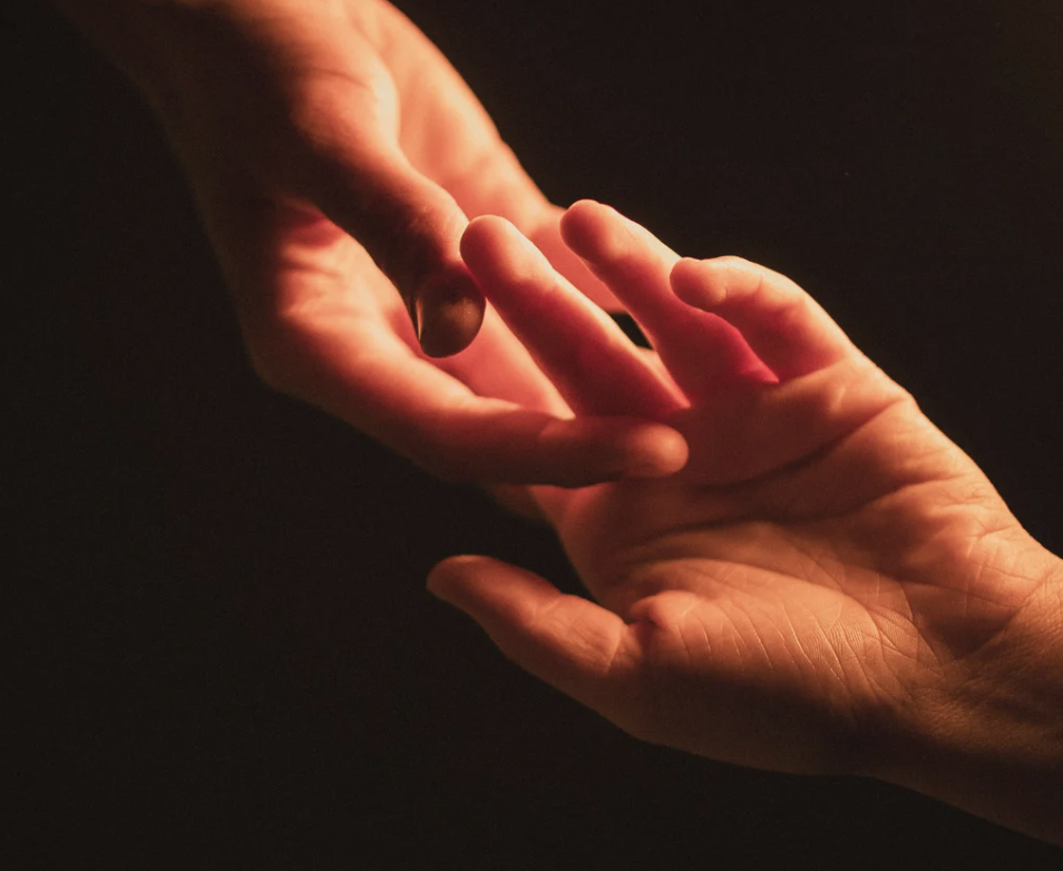 Close-up of two hands reaching towards each other in a dimly lit setting, with warm light illuminating their fingers.