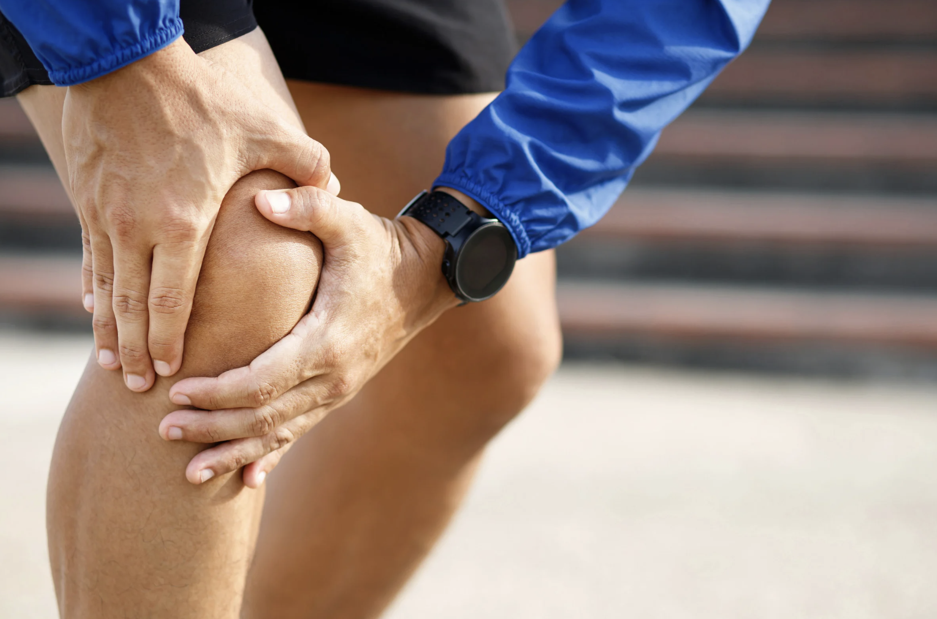 Person holding their knee in pain during exercise or running, wearing a blue jacket and sports watch.