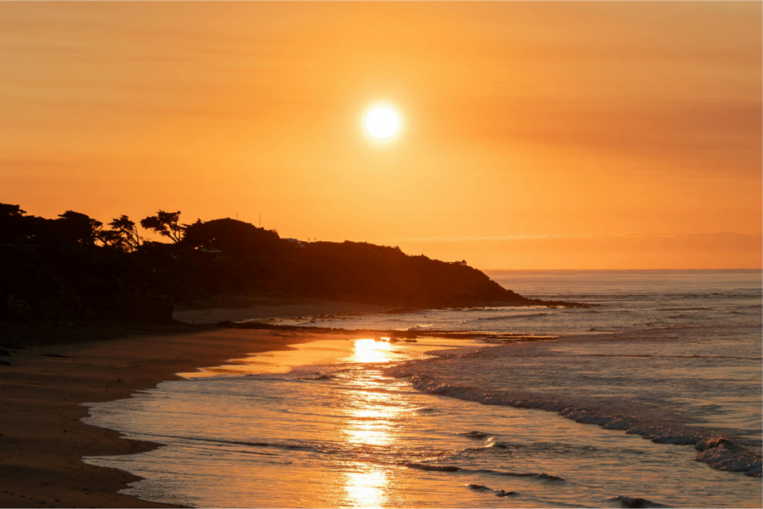 Sunset over a beach with waves and a silhouette of trees on a hill.