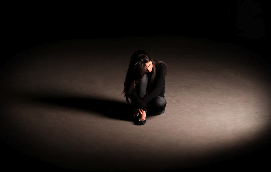 A woman sitting on the ground in a dark room, illuminated by a spotlight, with her head bowed and arms crossed over her knees, appearing distressed or contemplative.