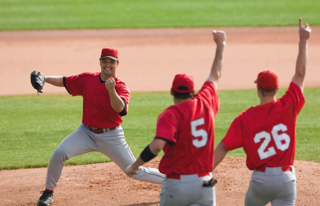 A baseball player in a red shirt and gray pants celebrating on a baseball field with two teammates, both in red jerseys, giving high-fives.