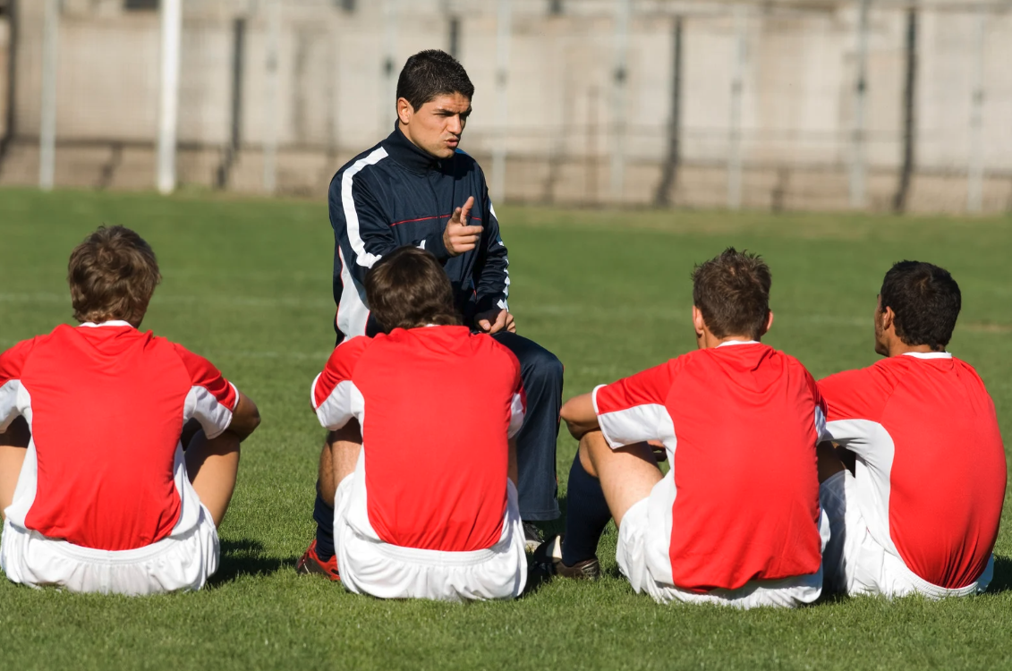A coach giving instructions to a soccer team sitting on the grass field.