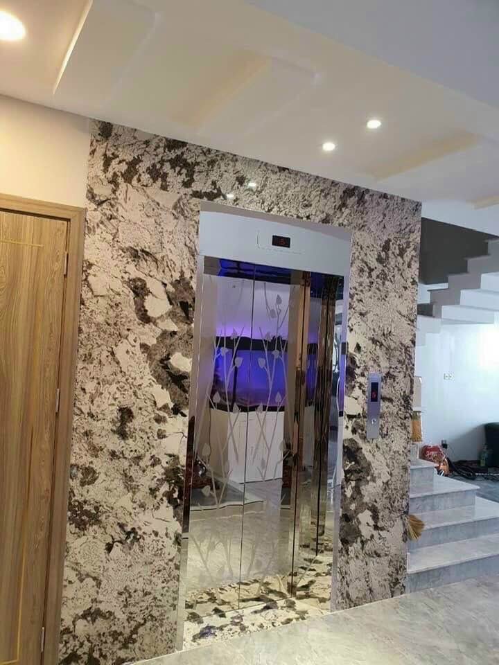 Modern home interior featuring an elevator with glass doors and floral patterns, surrounded by granite walls. Wooden door on the left and a staircase on the right.