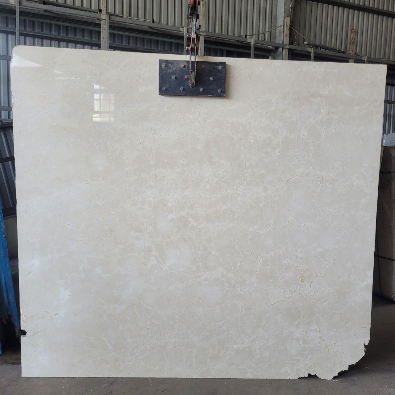 Large beige marble slab with textured surface in a warehouse setting, hanging from metal hooks.