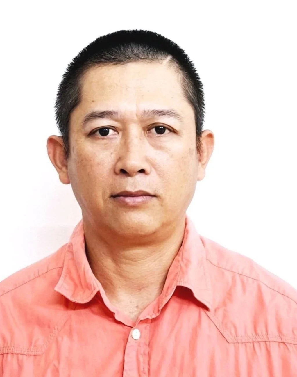 A portrait of a production manager with short black hair, wearing a coral-colored button-up shirt, facing forward against a plain white background.