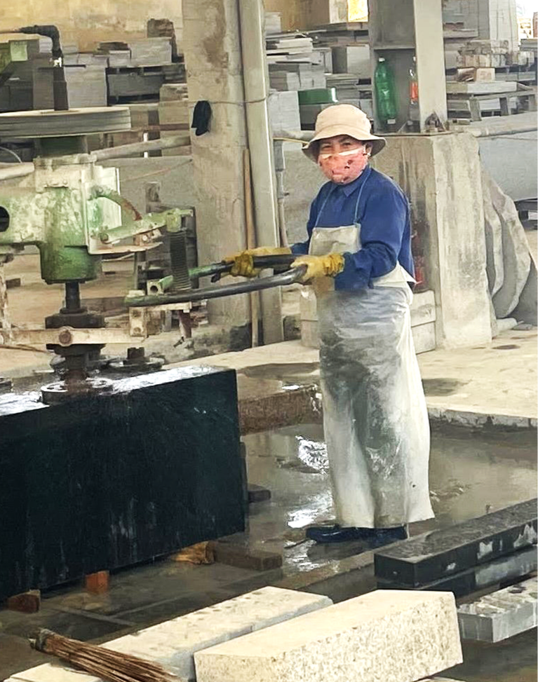A Vietnamese worker in a factory or workshop using a large green machine to cut or shape a black stone slab. The worker is wearing a beige hat, face mask, gloves, and an apron.