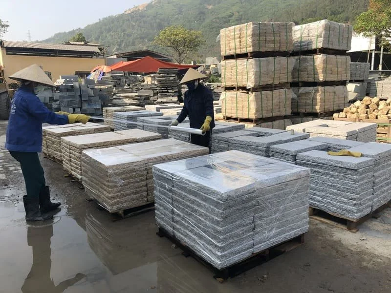 Workers in a stone yard stacking granite slabs with wrapped edges, wearing traditional conical hats and gloves; stacks of wrapped stone tiles in the background.