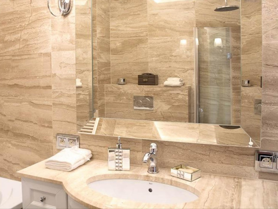 Modern bathroom with beige marble surfaces, a large mirror, a round sink, chrome faucet, folded towels, and a glass shower enclosure.