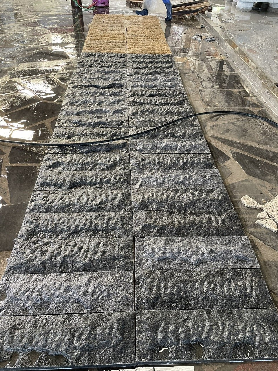 Textured stone tiles laid out in a long row on a wet floor, with some construction debris around.