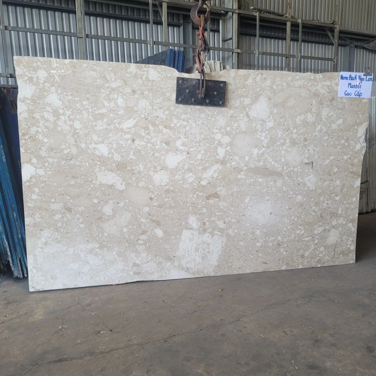 Large beige marble slab suspended by a hook in an industrial warehouse setting.