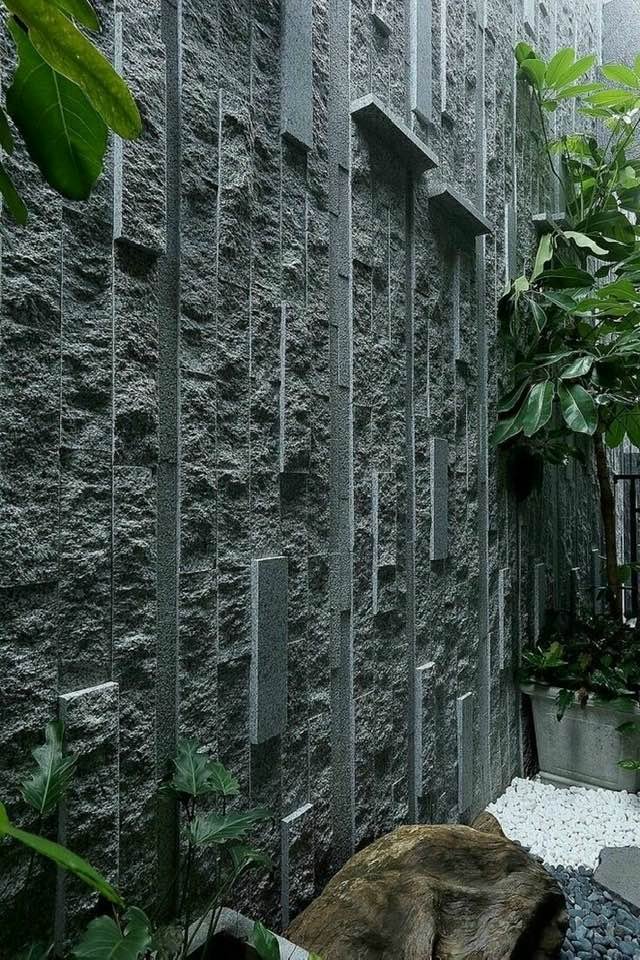 Vertical stone wall with textured geometric patterns, surrounded by green potted plants, pebbles, and a wooden log.