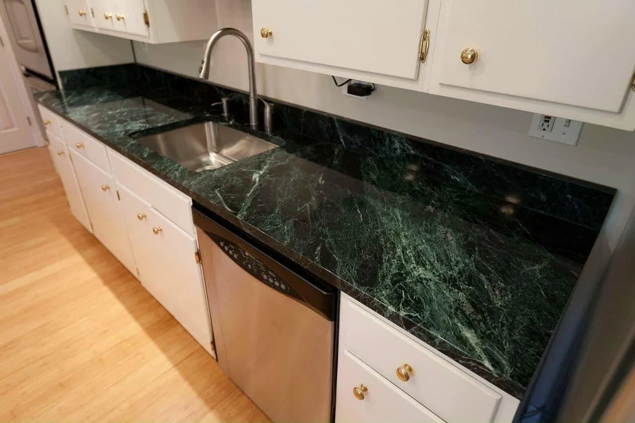 Modern kitchen countertop with dark green marble, stainless steel sink, white cabinets with gold knobs, and a dishwasher.