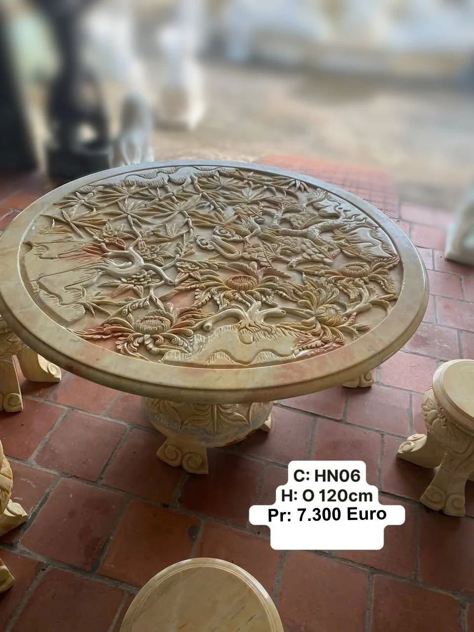 Round marble table with intricate carved design of trees and plants, supported by a decorative base, placed on a brick floor with other sculptures around.