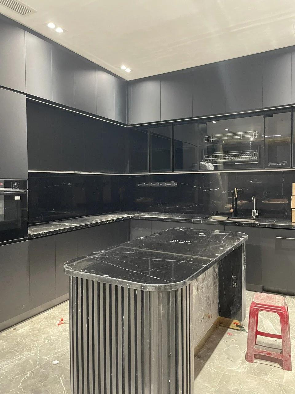 Modern kitchen interior with black cabinets and countertop, L-shaped layout, built-in oven, glass-fronted upper cabinets, and a red plastic stool.