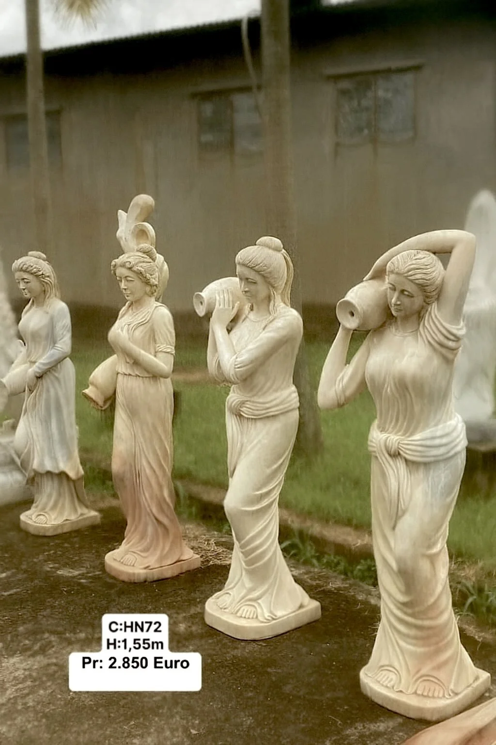 Row of four cream-colored concrete statues of women, each carrying a large vessel on their shoulder or in their hands, standing outdoors on grass.