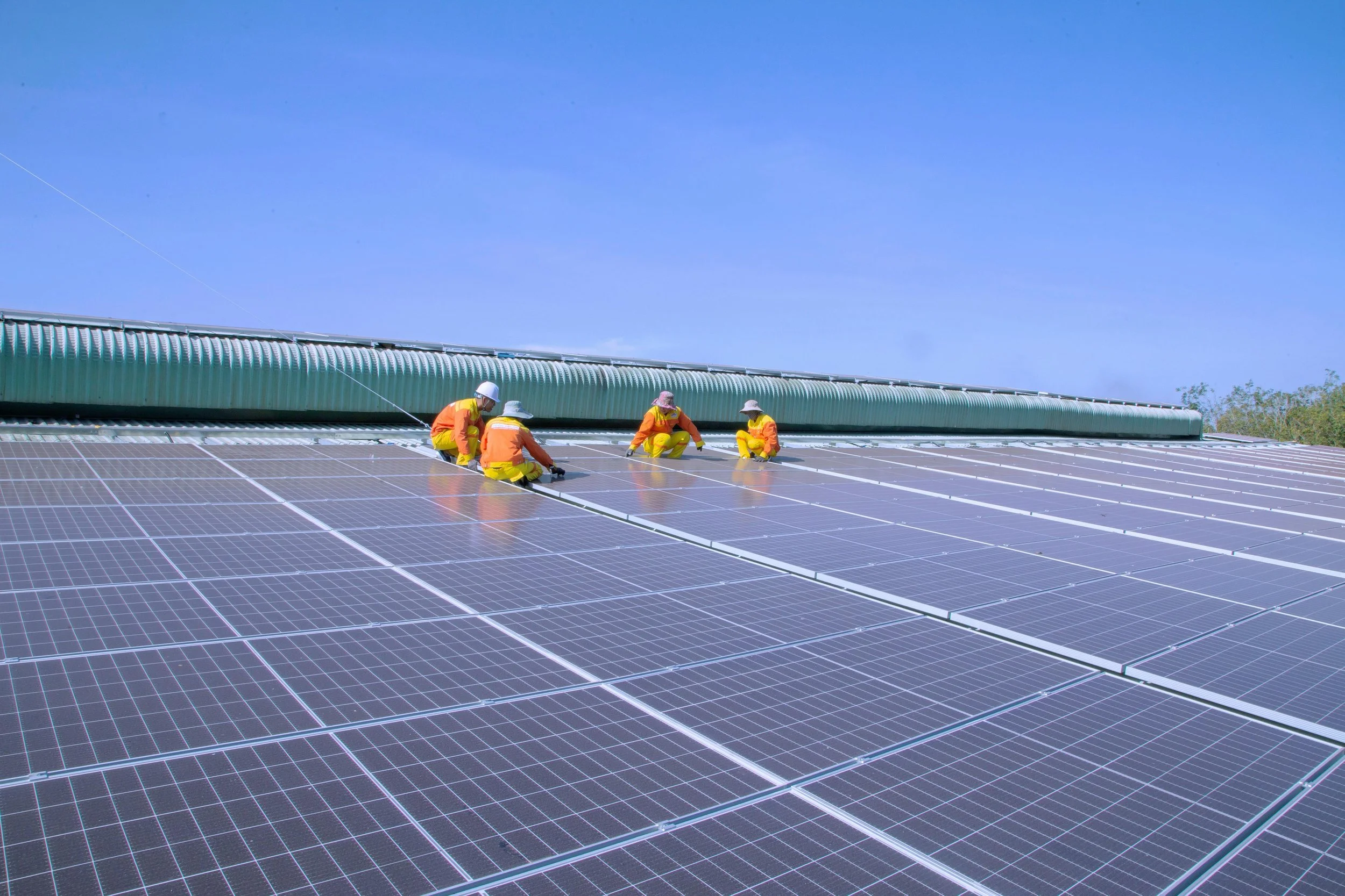 Workers in safety gear installing or maintaining solar panels on a rooftop against a bright blue sky.