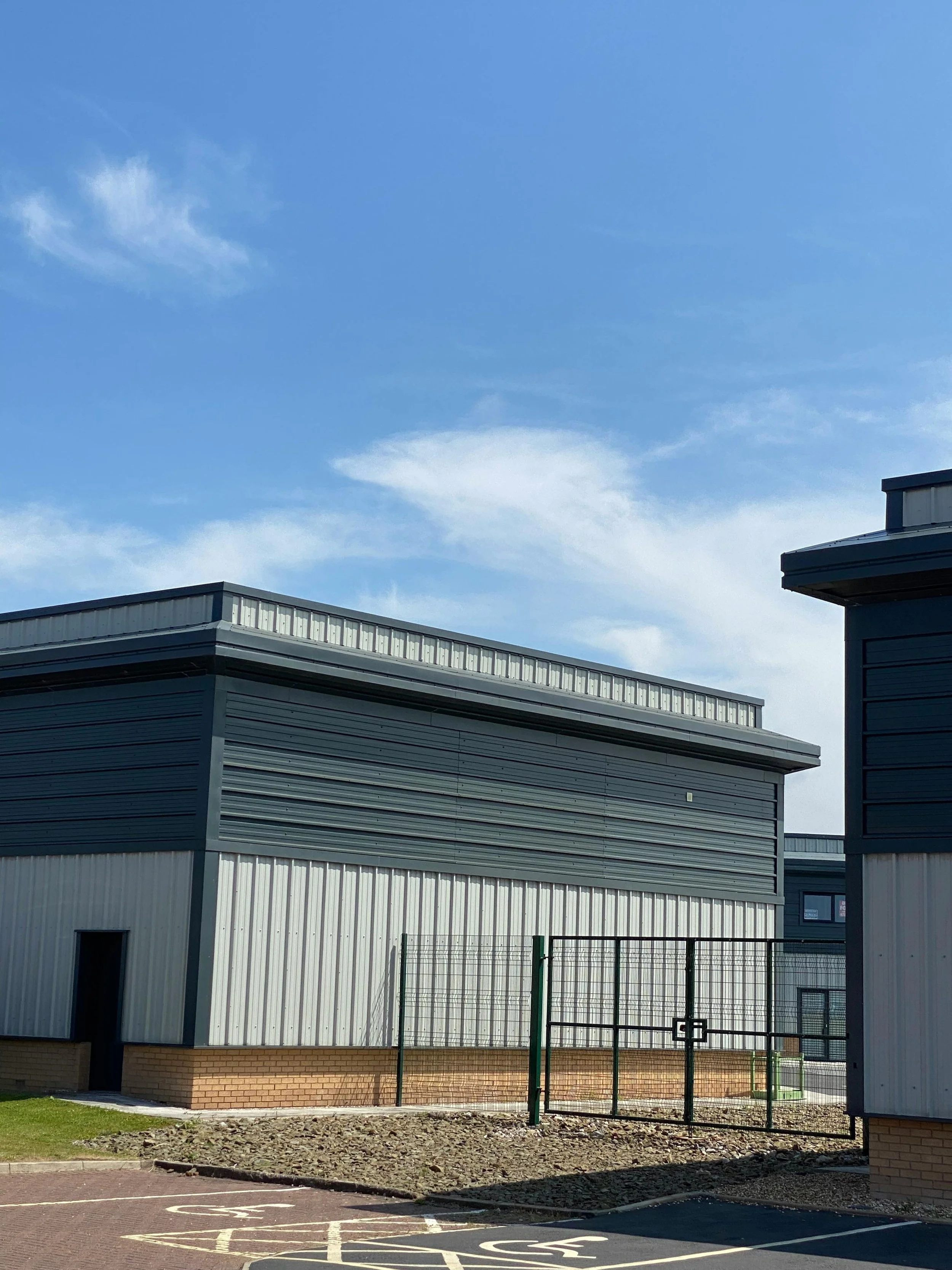 Industrial building with metal siding, a fenced area, and nearby parking lot under a blue sky with clouds.