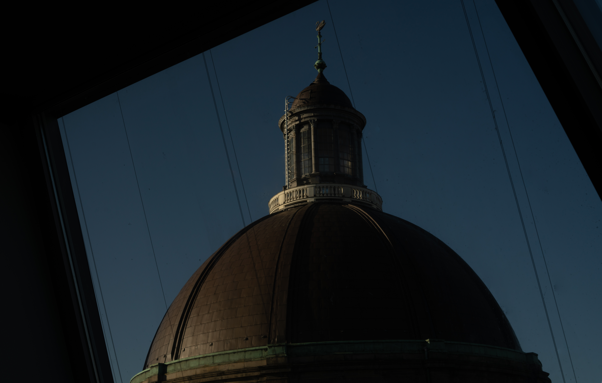 View of a building's dome with a small tower and weather vane, seen through a slanted window with visible window frame and wires in the background