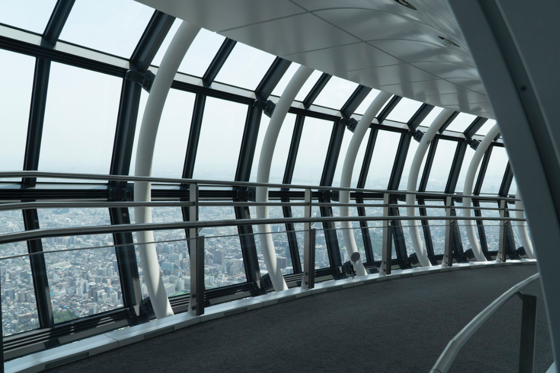 Interior view of a futuristic observation deck with large glass windows, cityscape view, curved white structural beams, and metal railings.