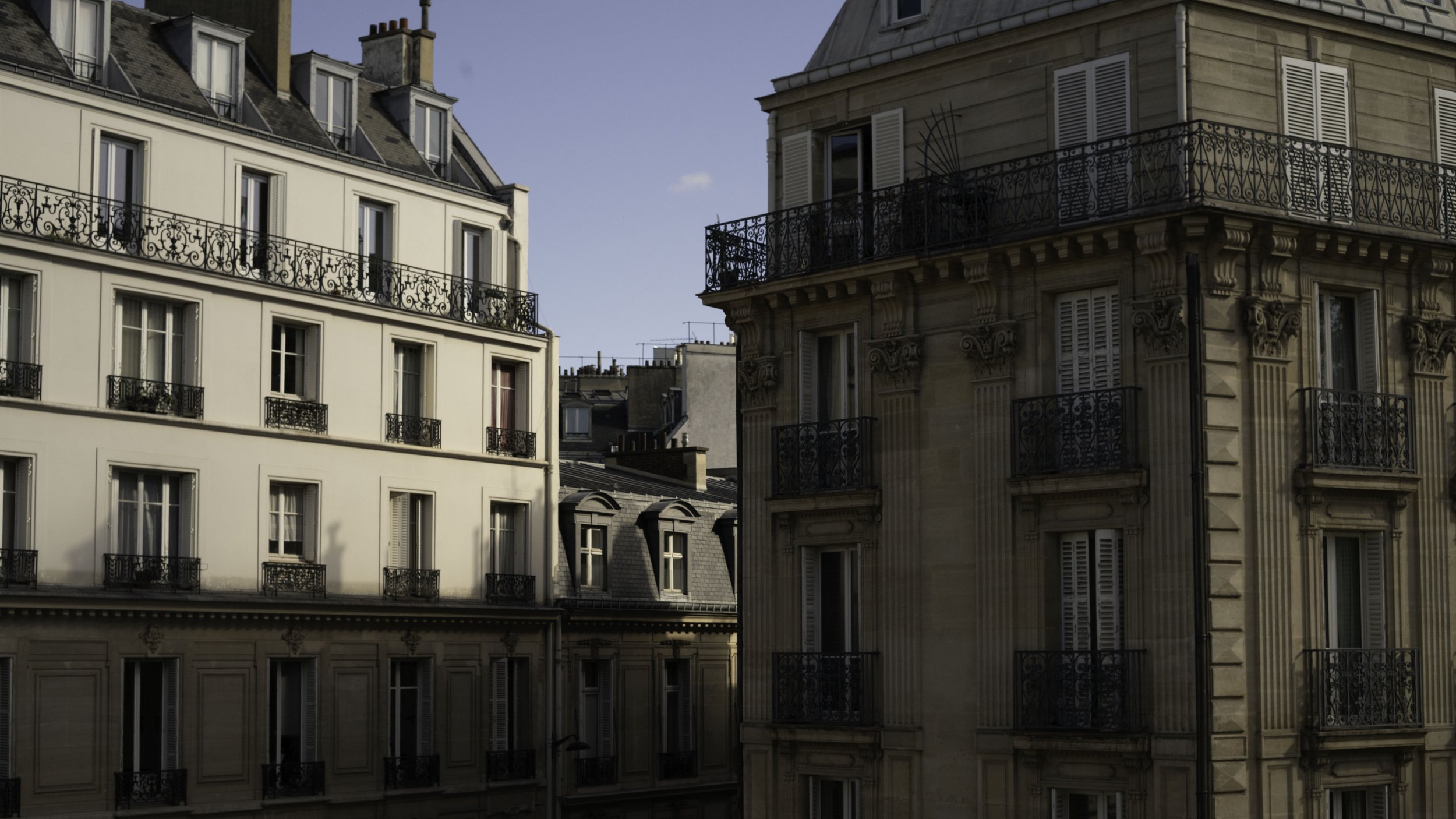 Parisian apartment buildings with detailed architecture, black wrought-iron balconies, and grey slate roofs under a partly cloudy sky