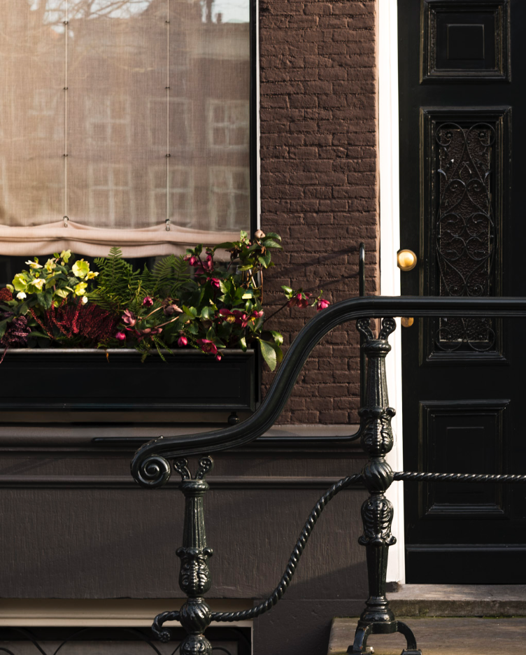 View of a house front with black door, brick wall, window with curtain, planter box with flowers, and decorative black railing.