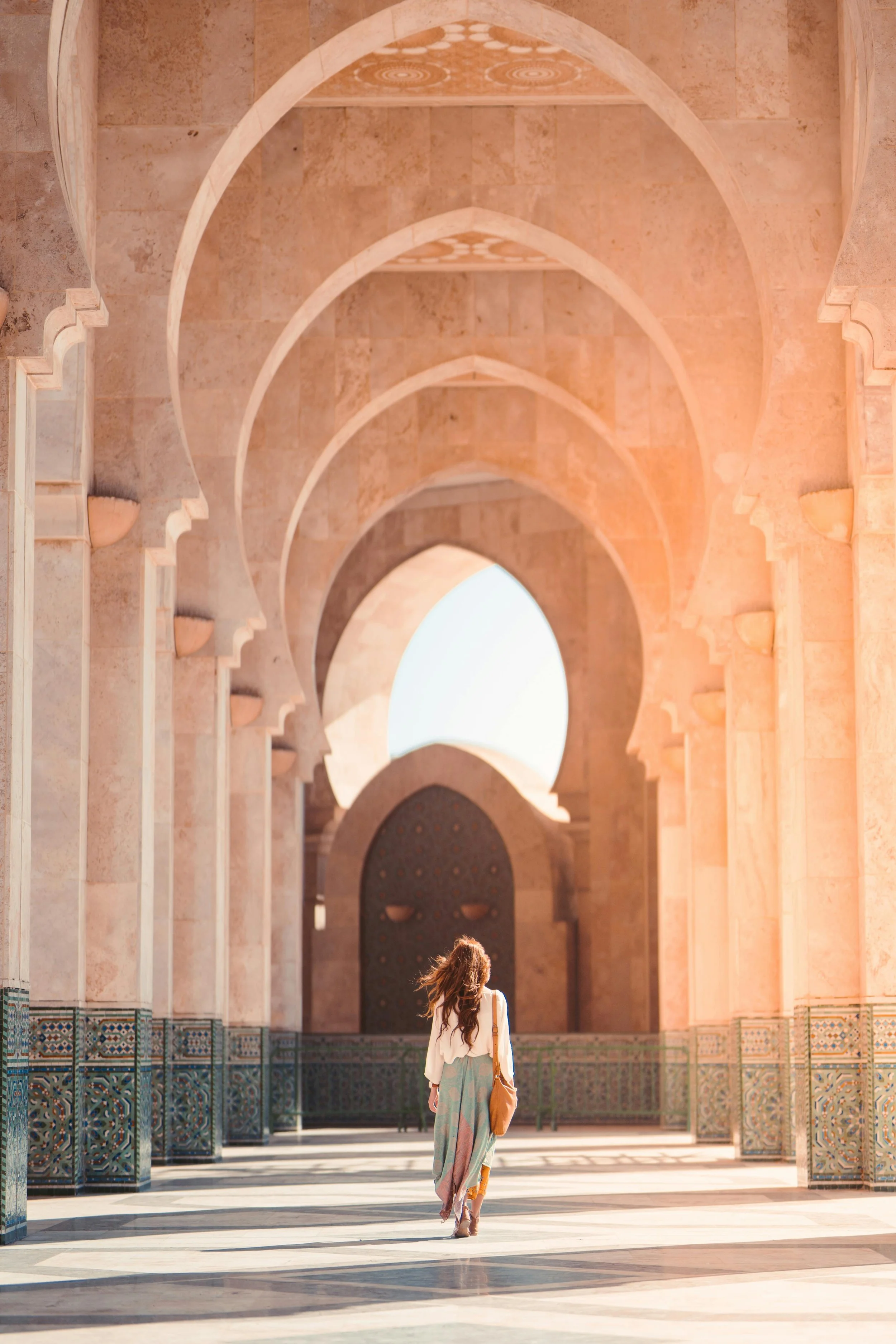 Woman in long dress walking through an ornate stone archway, with intricate tilework and soft natural lighting.