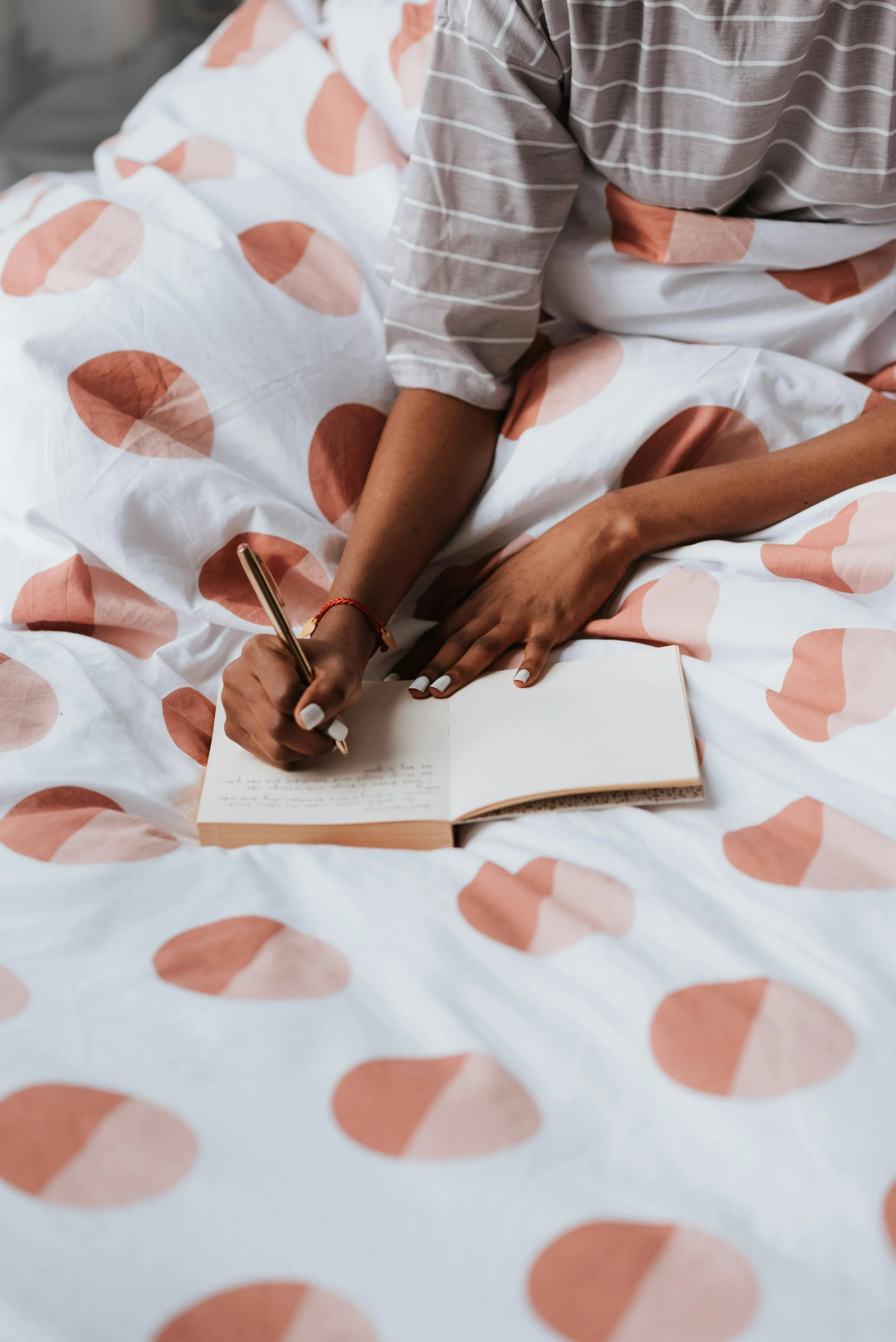 Person writing in a journal on a bed with white and pink polka dot sheets.