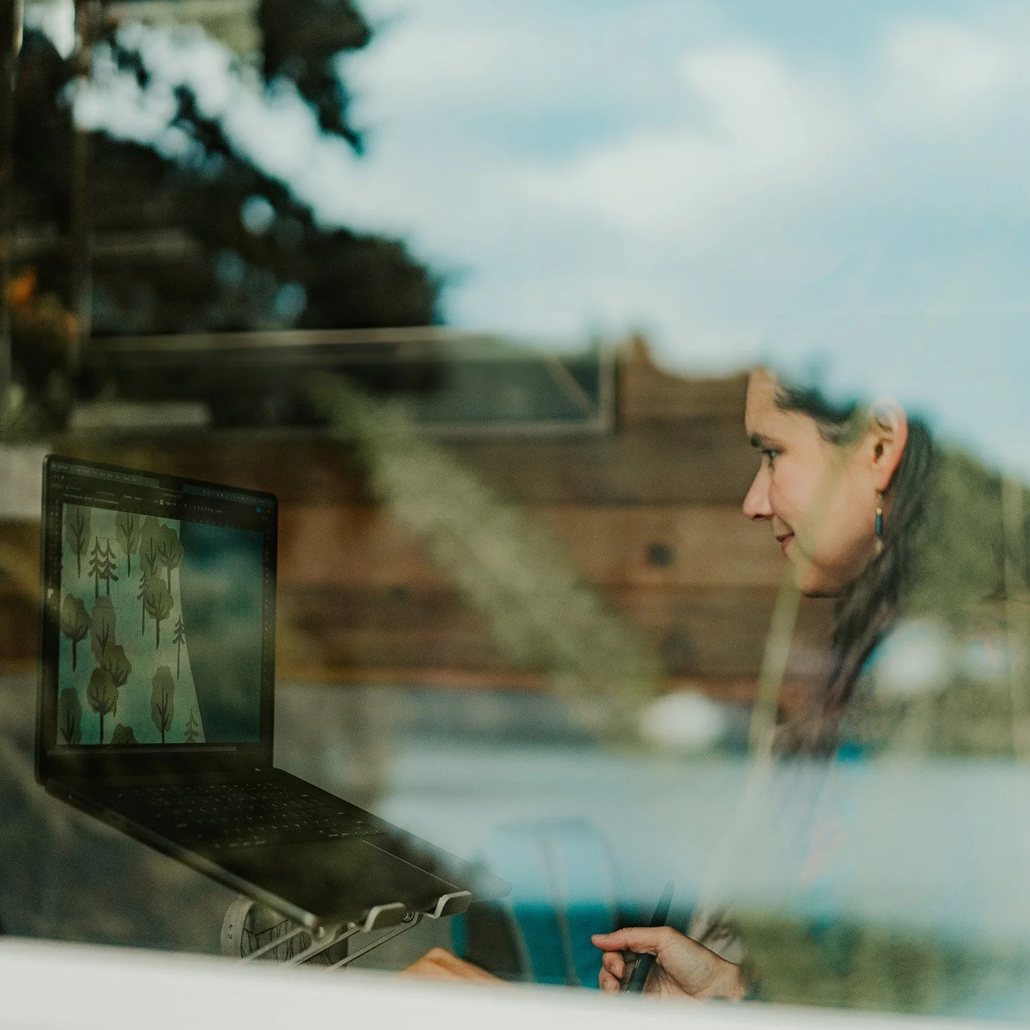 Trina is seen working on a laptop, through a window, with reflections of nature seen in the window glare.
