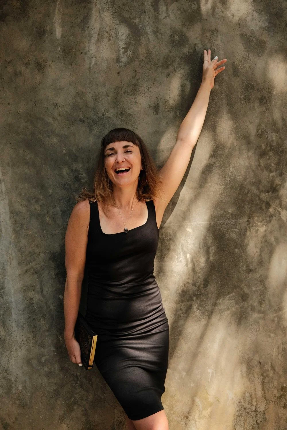 Dr Stephanie Minchin laughing, leaning against a textured stone wall in dappled light