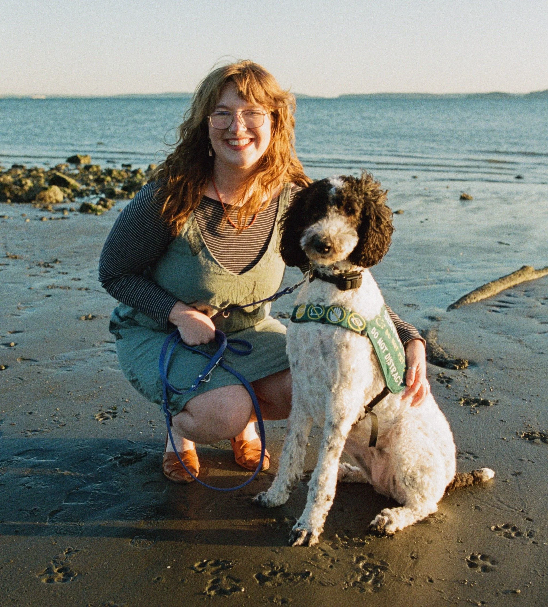 Anna, a blonde white woman with bangs and glasses, squats next to a black and white parti poodle wearing a service dog vest. They are on the beach and you can see sand, rocks and ocean behind them.
