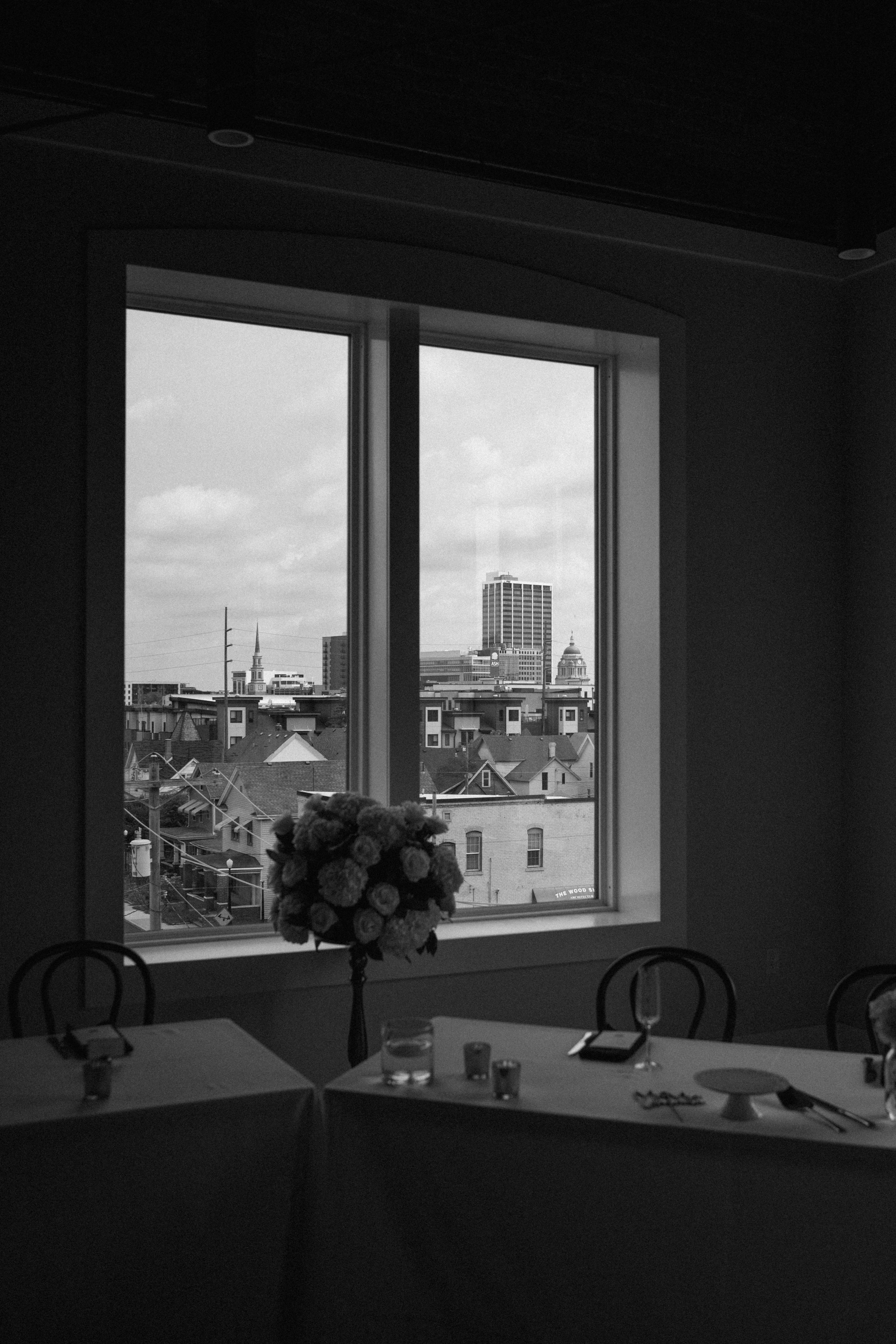 A table with tableware and a large flower arrangement inside a room, overlooking a city skyline through a window.