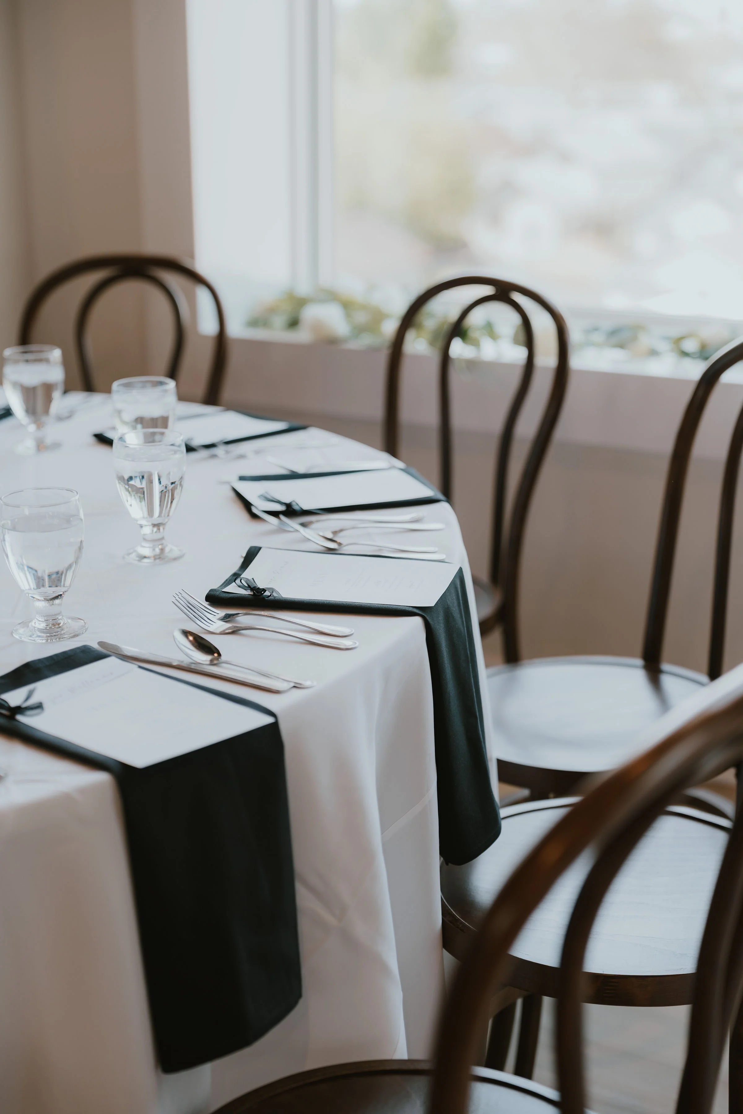 Elegantly set round dining table with white tablecloth and black table runner, four glasses of water, silverware, and black napkins in a well-lit room with large window and wooden chairs.